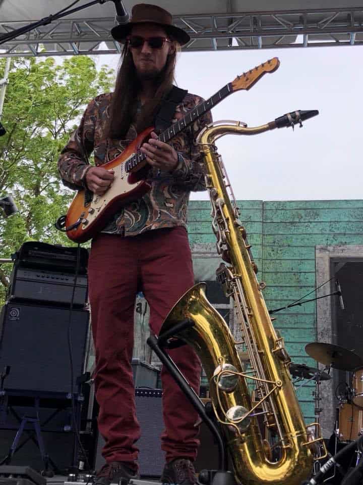 Picture of Brandon Logan playing his guitar, with his saxophone sitting on a stand in front of him, on stage at the Journey To Memphis band competition - at the Portland Blues Festival, at the Portland Oregon Waterfront.