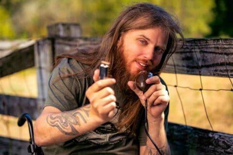 Johnny Wheels in front of a fence on his farm, talking into a microphone in his left hand and pointing to "you" with the harmonica in his right hand.
